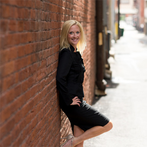 Monica Kerber leaning against a brick wall in a black skirt suit.